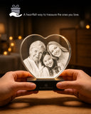 A heart-shaped clear crystal photo plaque on a black base, held by two hands in a warmly lit room. The crystal shows a grayscale portrait of three women, with blurred warm lights in the background.