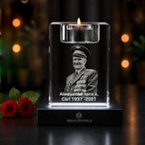 A clear acrylic memorial candle holder with a lit tealight on a black base, engraved with a black-and-white portrait and the text 'Always our hero Carl 1937-2021.' Two red roses sit to the left on a dark surface against a softly blurred warm background.