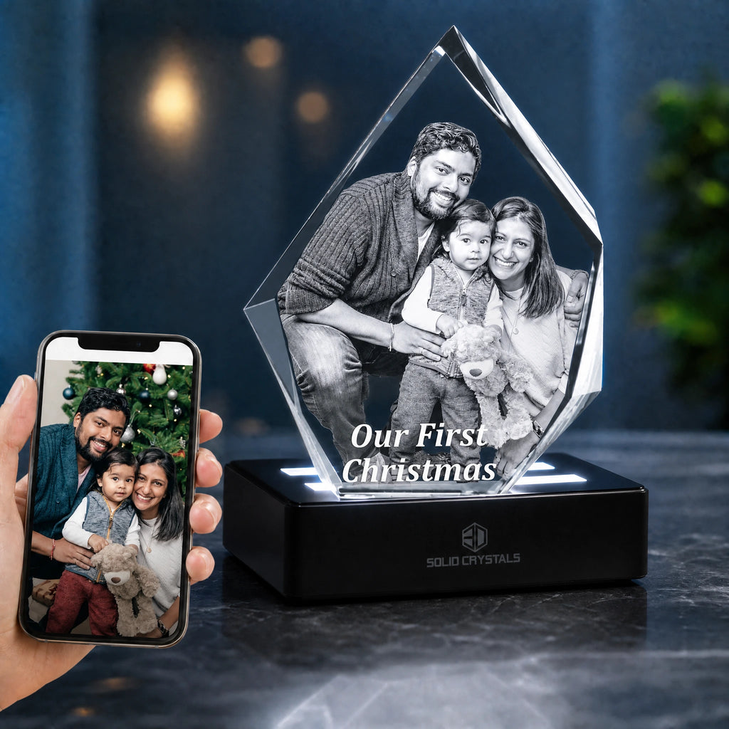 A personalized acrylic crystal award shaped like a house, featuring a printed family photo and the text 'Our First Christmas,' displayed on a black LED base. A hand holds a smartphone showing the original photo on the left, with a dark, softly blurred indoor background. Gift shop, Personalised gifts, gift for her, gift for him, gift for pet, christmas gift, xmas, gift for her, gift for him, heart crystal, crystal heart, crystal 3d, crystal photo, corporate gift, corporate gift uk, 3DCrystal London, Gift sho