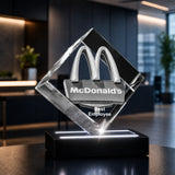 A clear acrylic award trophy shaped like a tilted square with the McDonald's golden arches logo at the top and the text 'Best Employee' on the front, displayed on a black illuminated base in a modern office lobby.